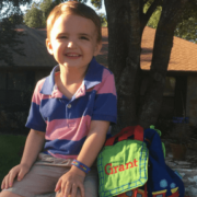Boy in blue and pink horizontal stripe shirt smiling outdoors with lunchbox